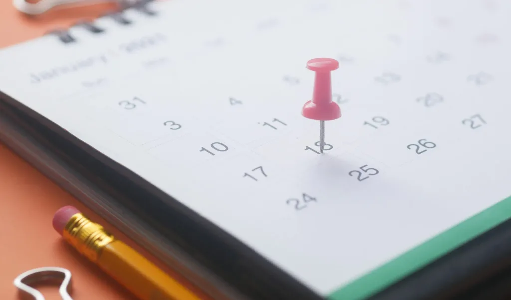 A close-up of a calendar page with a pink push pin, pencil, and binder clip on an orange background.